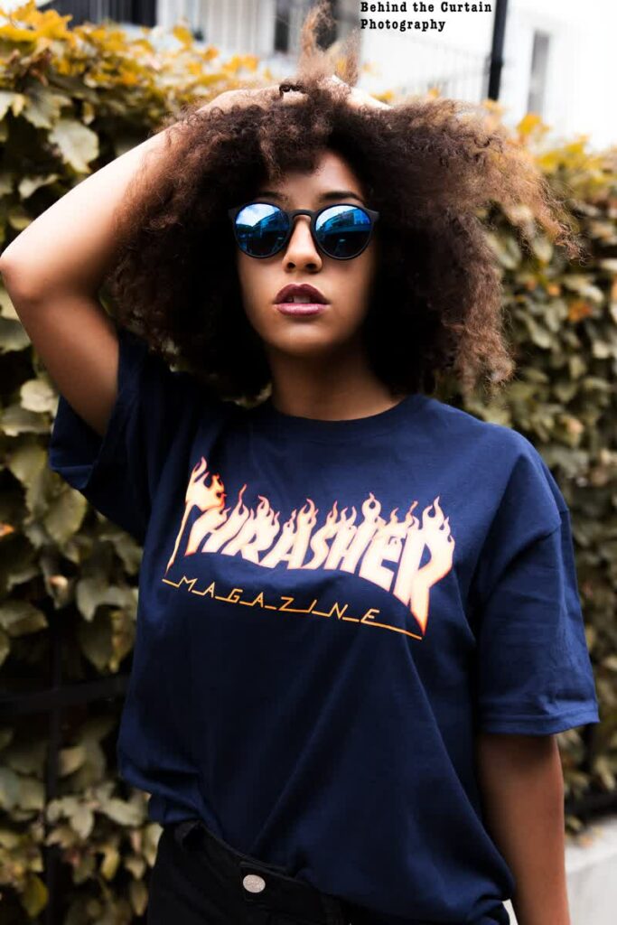 eine schwarze Frau mit lockigen Haare und einer Sonnenbrille auf im T-Shirt vor einer Hecke