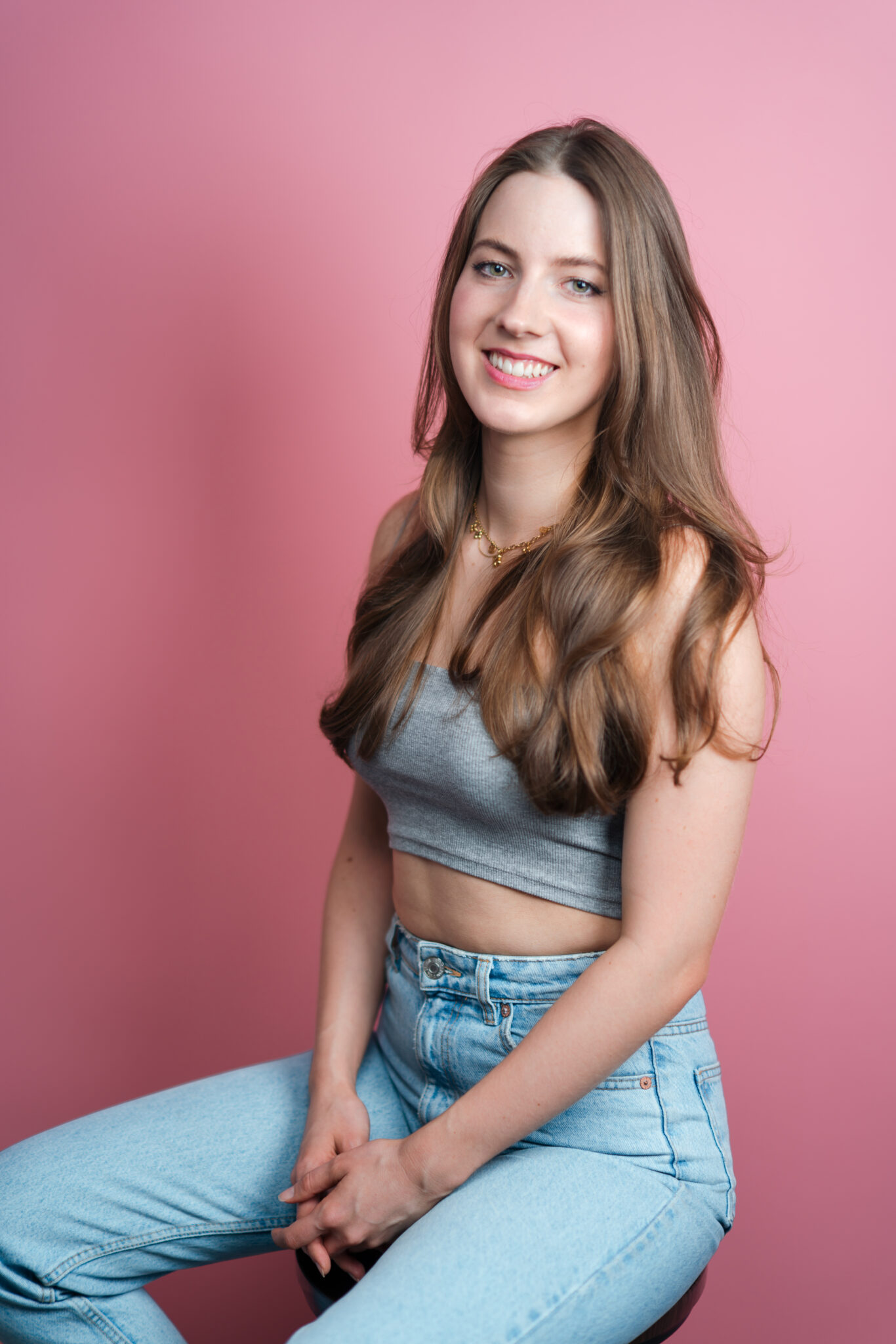 73R08246-3 Junge Frau sitzt im Fotostudio vor einem rosa Hintergrund auf einem Hocker und lächelt