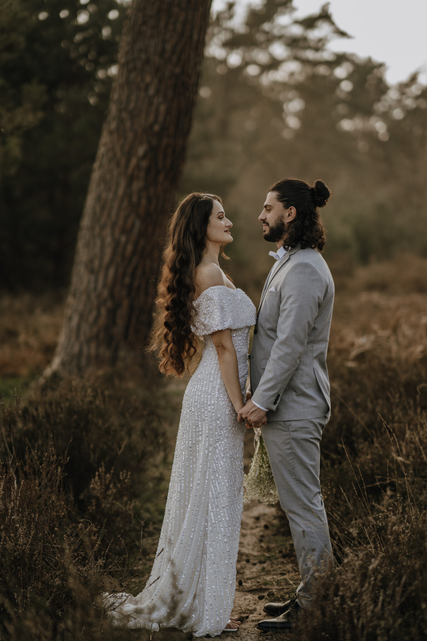 73R01284 Bei dem After Wedding Shooting stehen die Braut und der Bräutigam gegenüber voneinander. Sie halten sich gegenseitig die Hände, dabei sind die Arme von beiden nach unten ausgestreckt und sie gucken sich lächelnd in die Augen