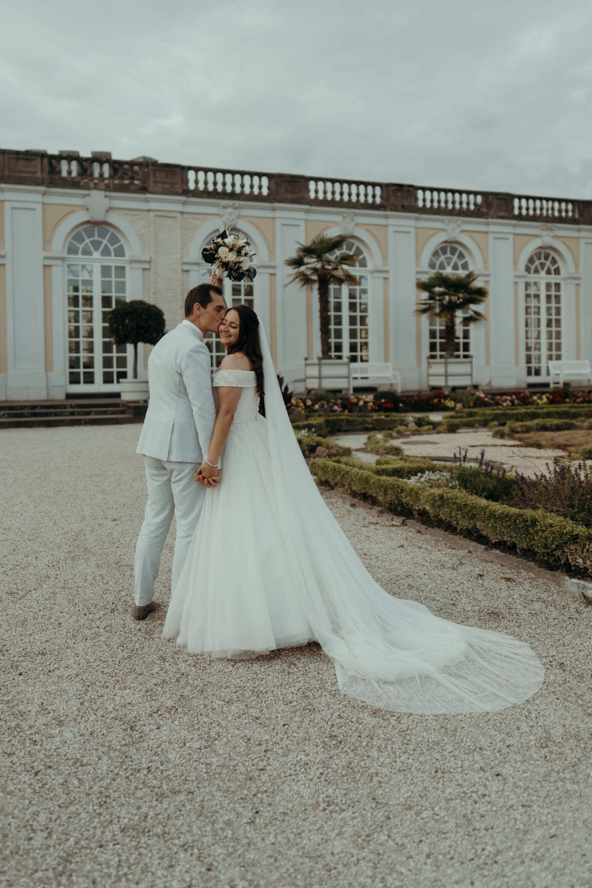 chrissandraafterwedding-6902-2 Kopie Hochzeitspaar in Schlossgarten, Bräutigam küsst Braut auf Wange, Braut hält Strauß hoch