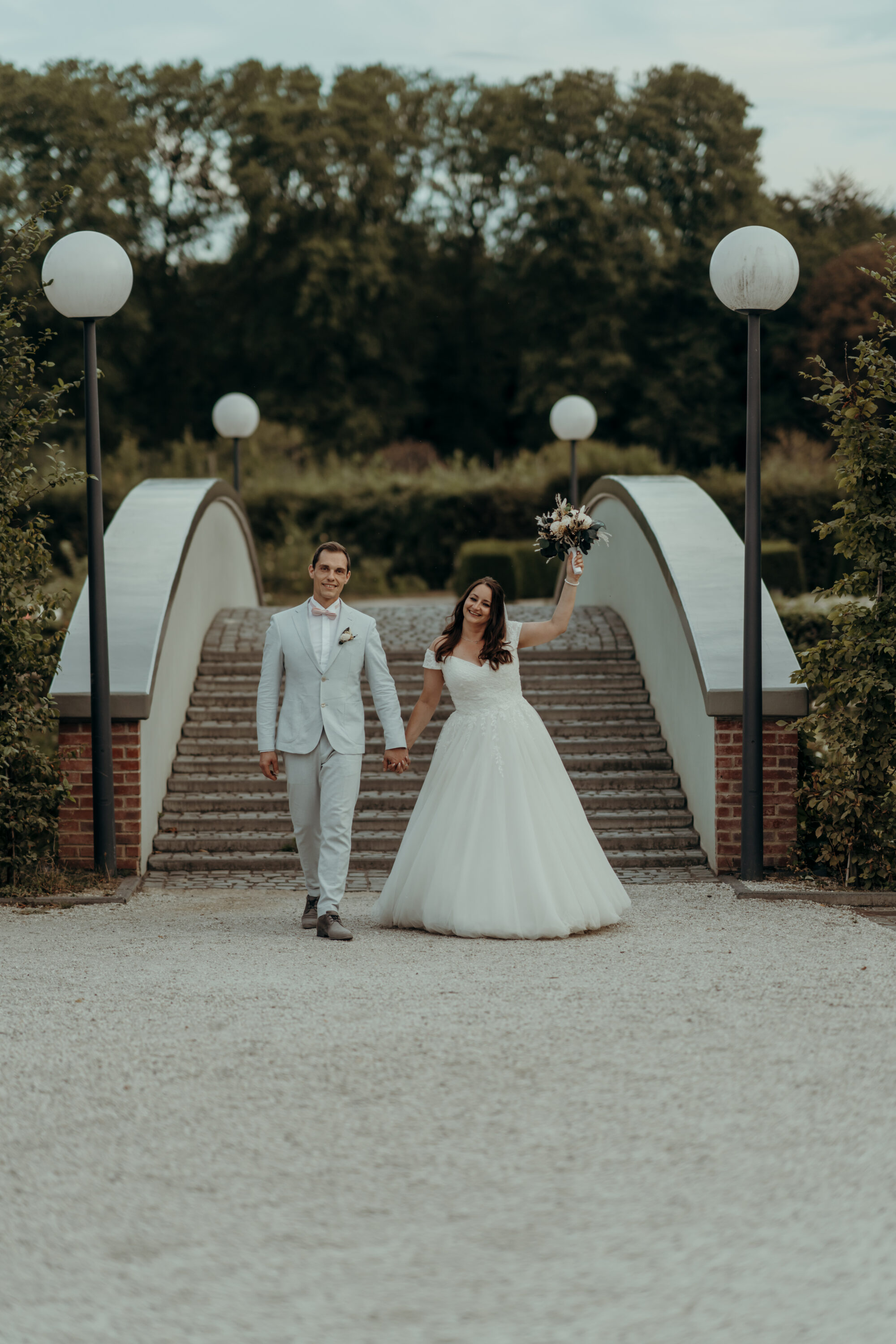 chrissandraafterwedding-7132-2 Kopie Brautpaar vor Brücke mit großen Laternen, auf Kamera zulaufend, Braut hält Strauß hoch