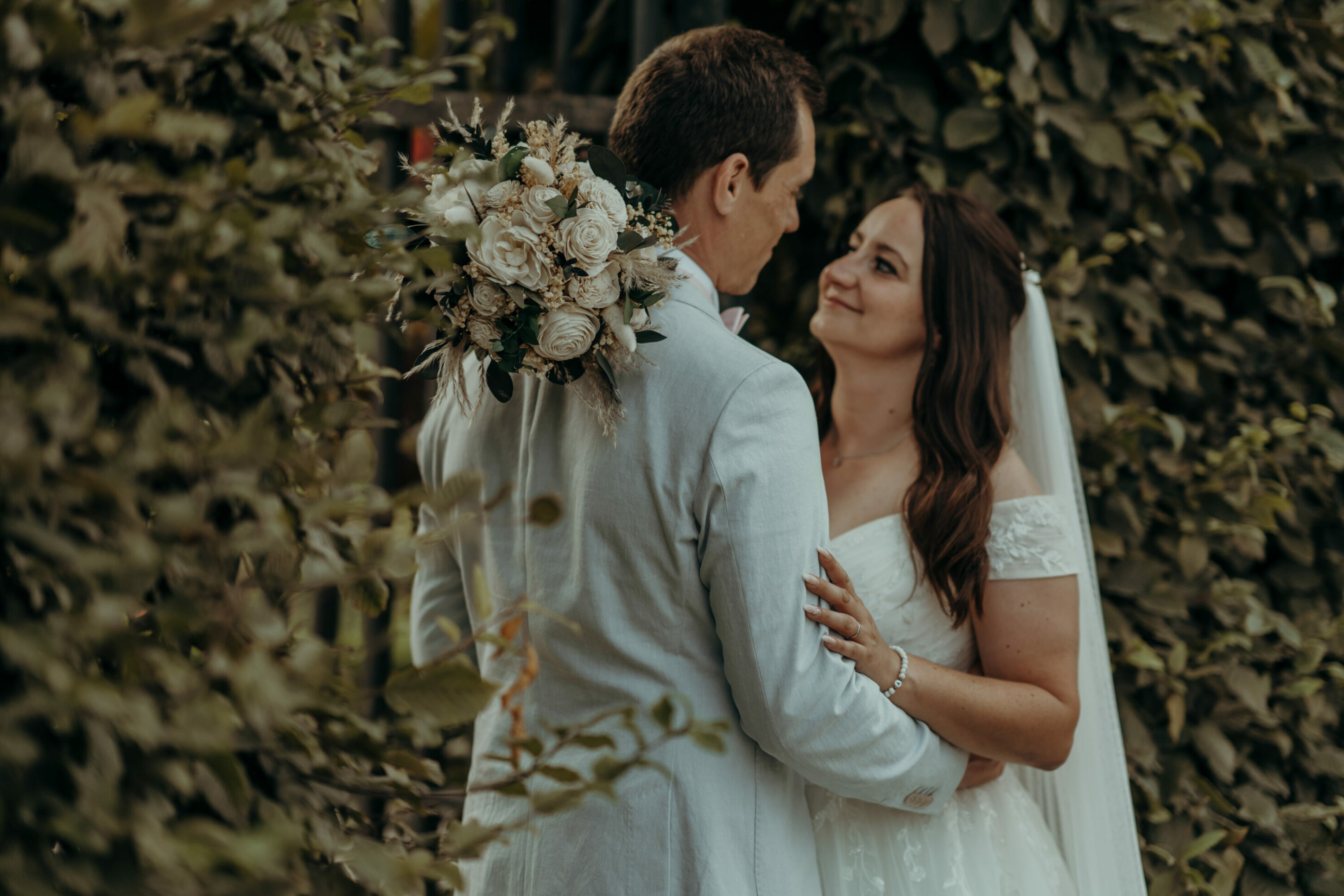 chrissandraafterwedding-7176-2 Kopie Brautpaar schaut sich in die Augen, gerahmt von Hecken, Brautstrauß fokussiert auf Schulter des Bräutigams