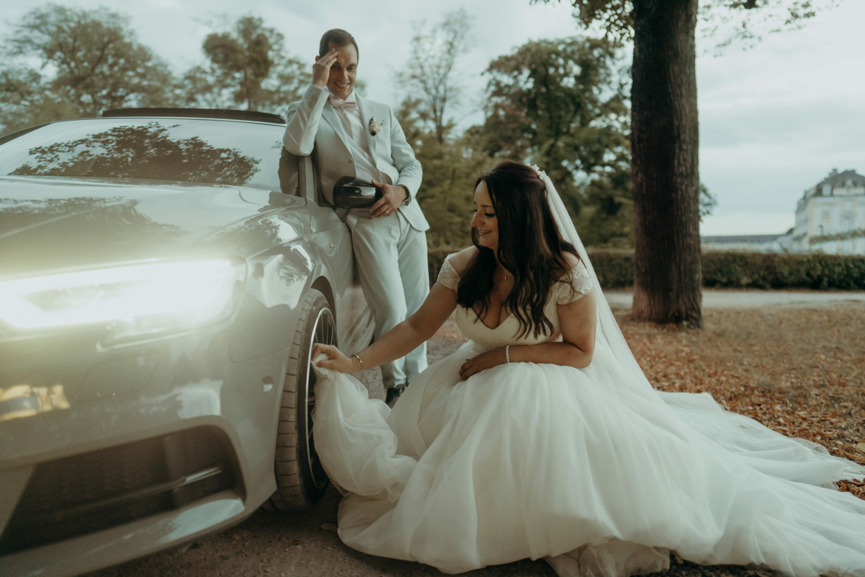 chrissandraafterwedding-7539-2 Kopie Braut putzt Felge des Autos mit Kleid, Bräutigam lacht und fasst sich an die Stirn