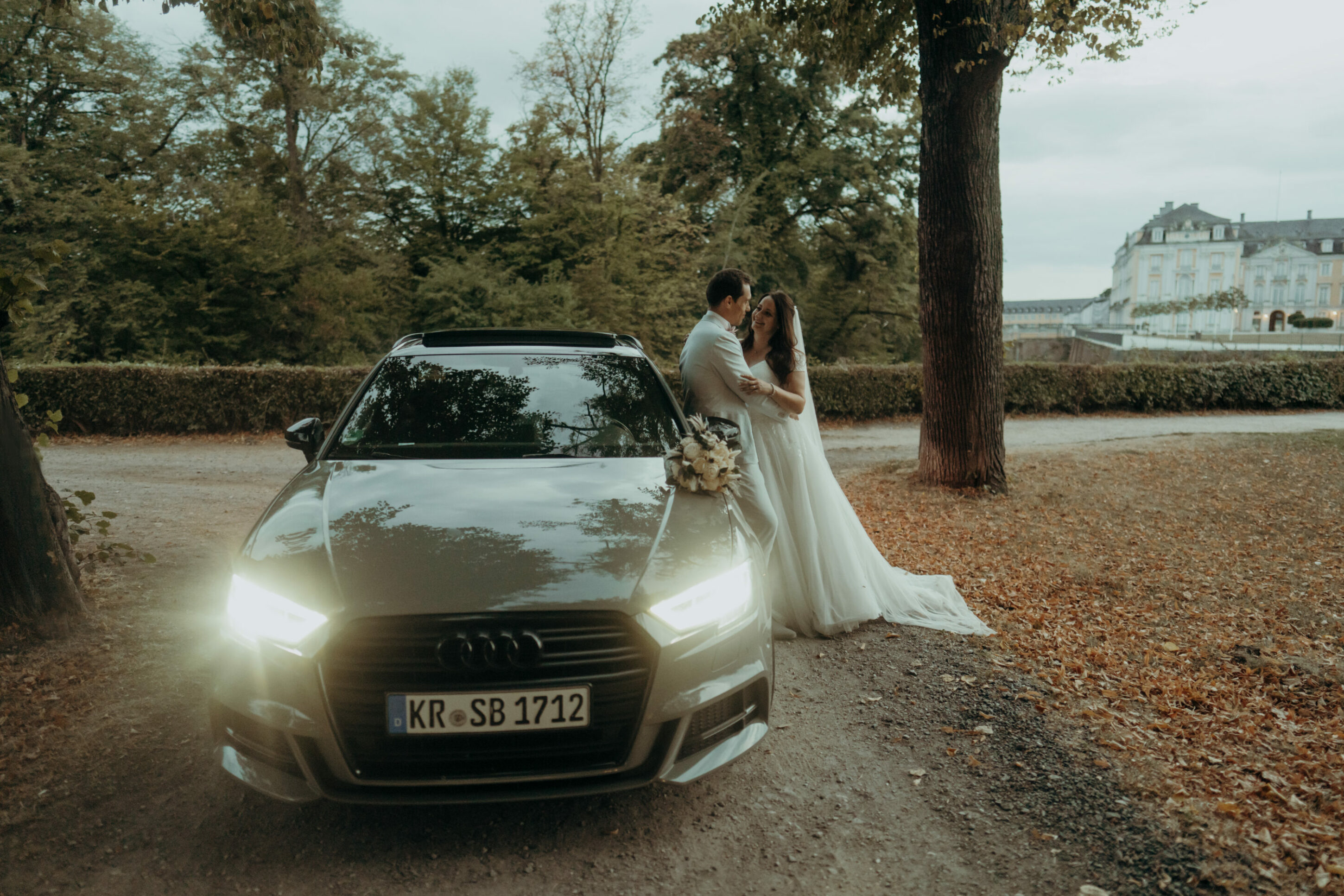 chrissandraafterwedding-7566-2 Kopie Hochzeitspaar lehnt an Audi, sich im Arm haltend, Schloss im Hintergrund, Allee