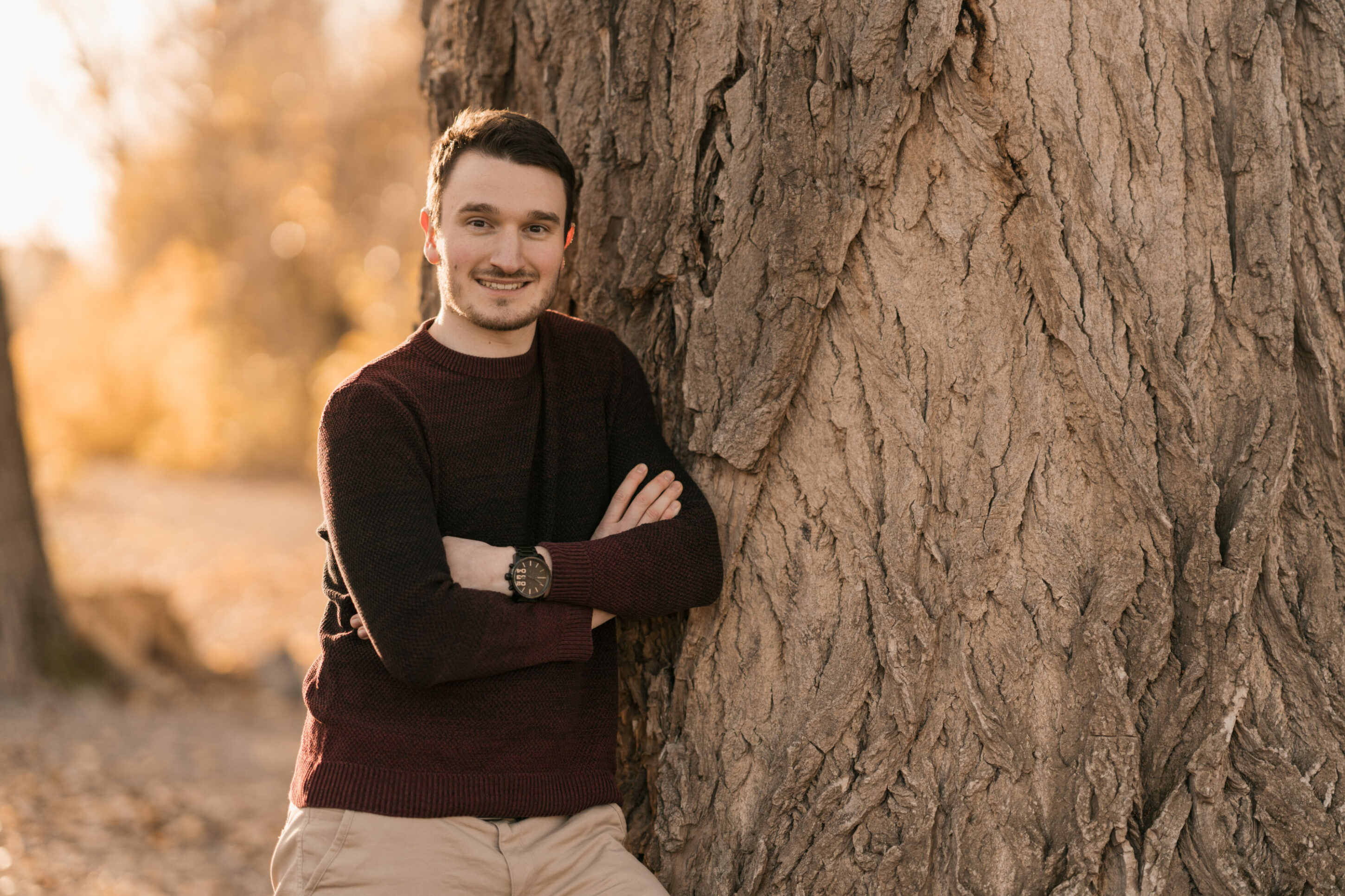 throg-3305 Junger Mann steht im Herbst an Baum gelehnt mit verschränkten Armen und lächelt