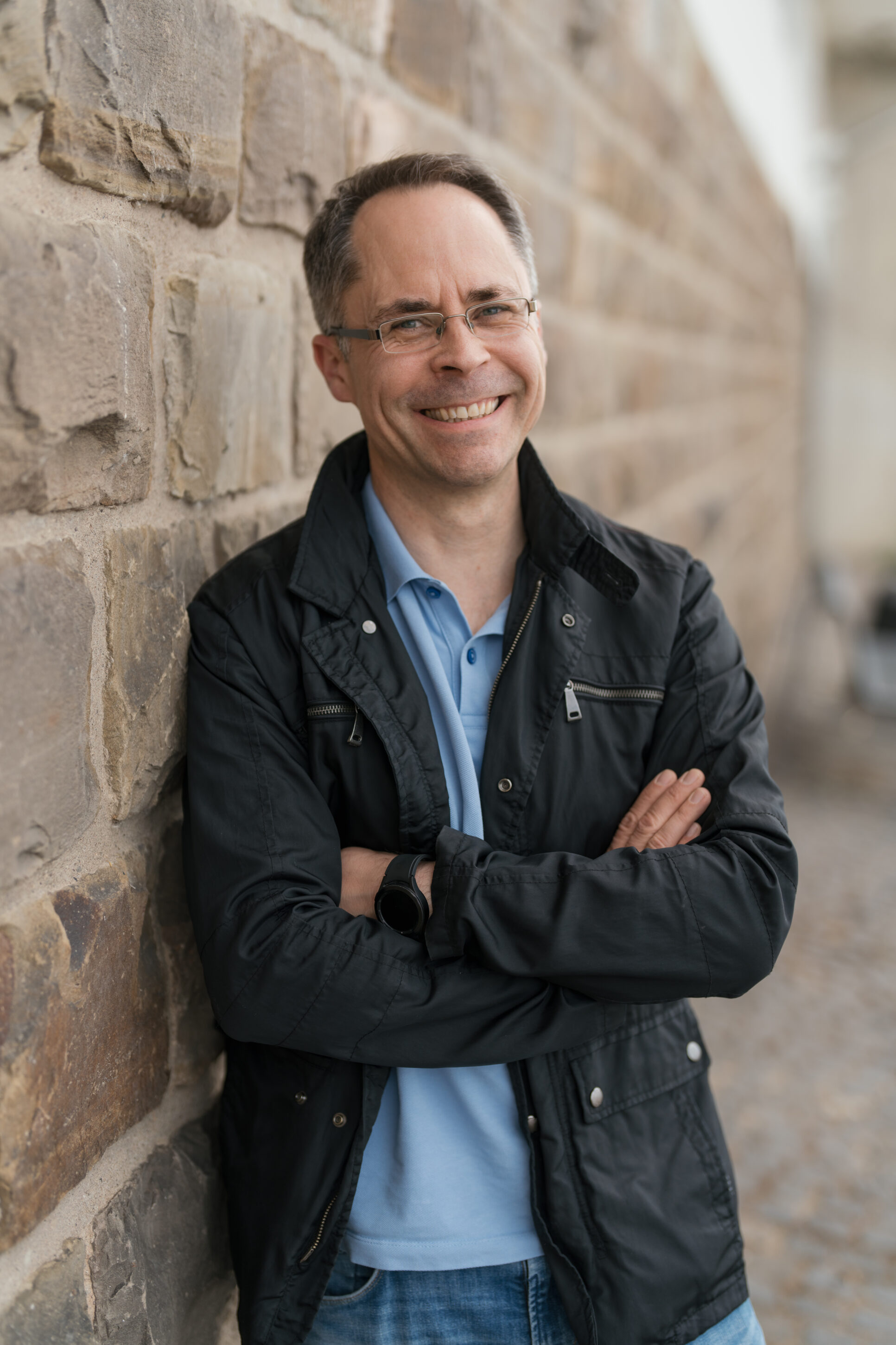 _4R17523-ma_sc_portrait_072023 Mann mit Brille bei Tinder Fotoshooting lehnt sich lächelnd an die Mauer