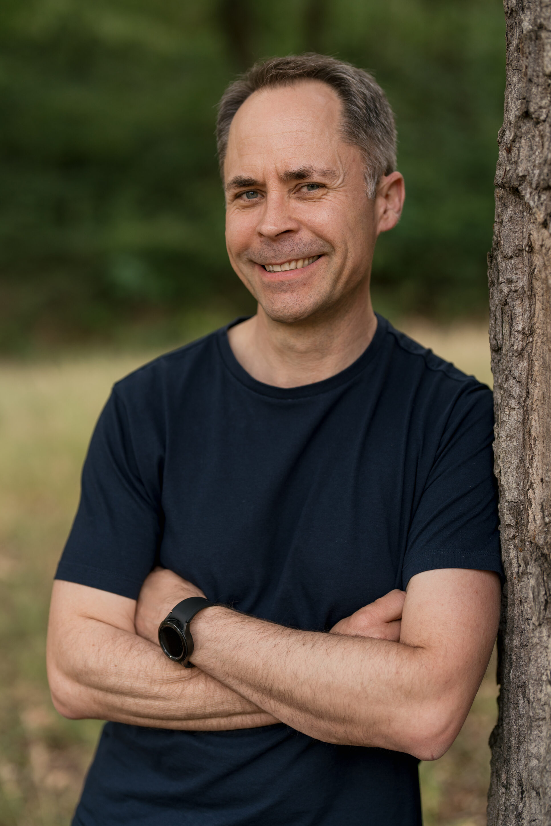 _4R18072-ma_sc_portrait_072023 Mann lehnt an einem Baum, schaut in die Kamera mit verschränkten Armen und lächelt dabei